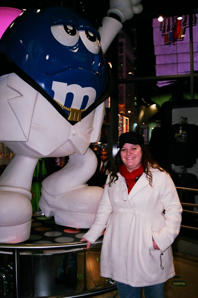 Me standing next to the blue M&M guy at the New York M&M store. Wearing a long cream jacket with a red scarf and black hat.