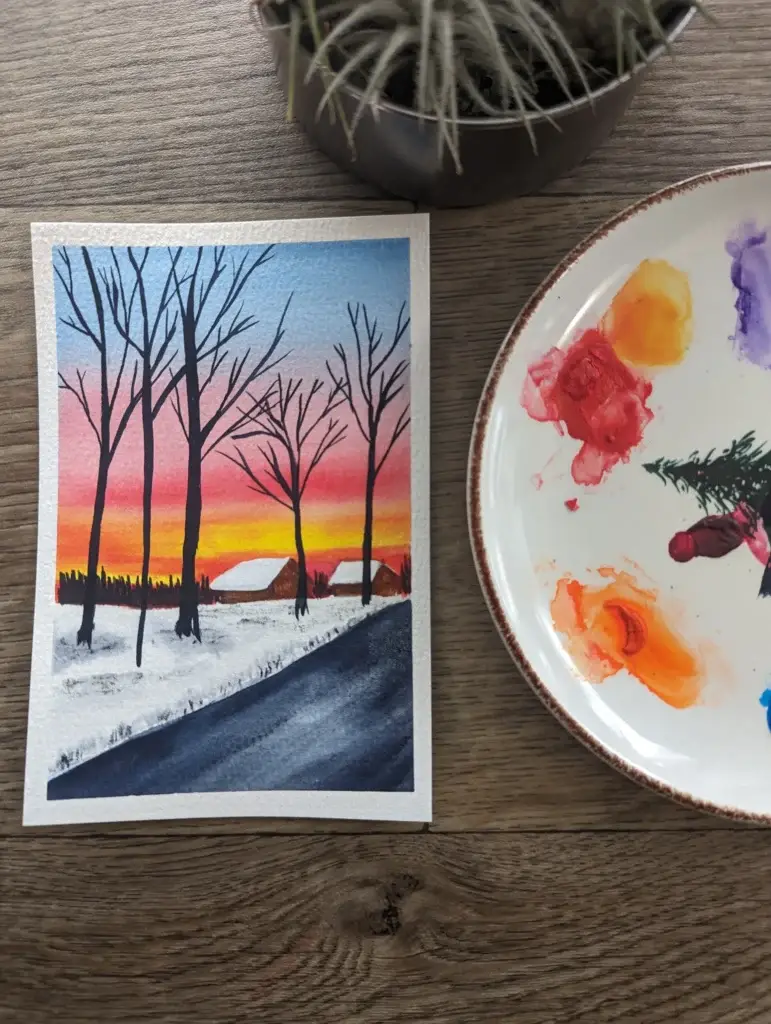 Watercolor painting of two small cabins on the side of the road. The foreground has black trees, with no leaves, just branches. The background has a sunset sky with light blue, red, and yellow mixing for a little bit of orange.