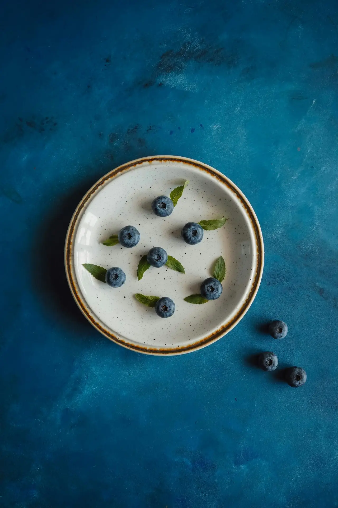 seven blueberries space out on a plate with leaves next to them.