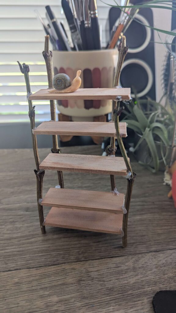 A miniature bookshelf project, made out of branches and a paint stirrer. On the top shelf there is a small clay snail