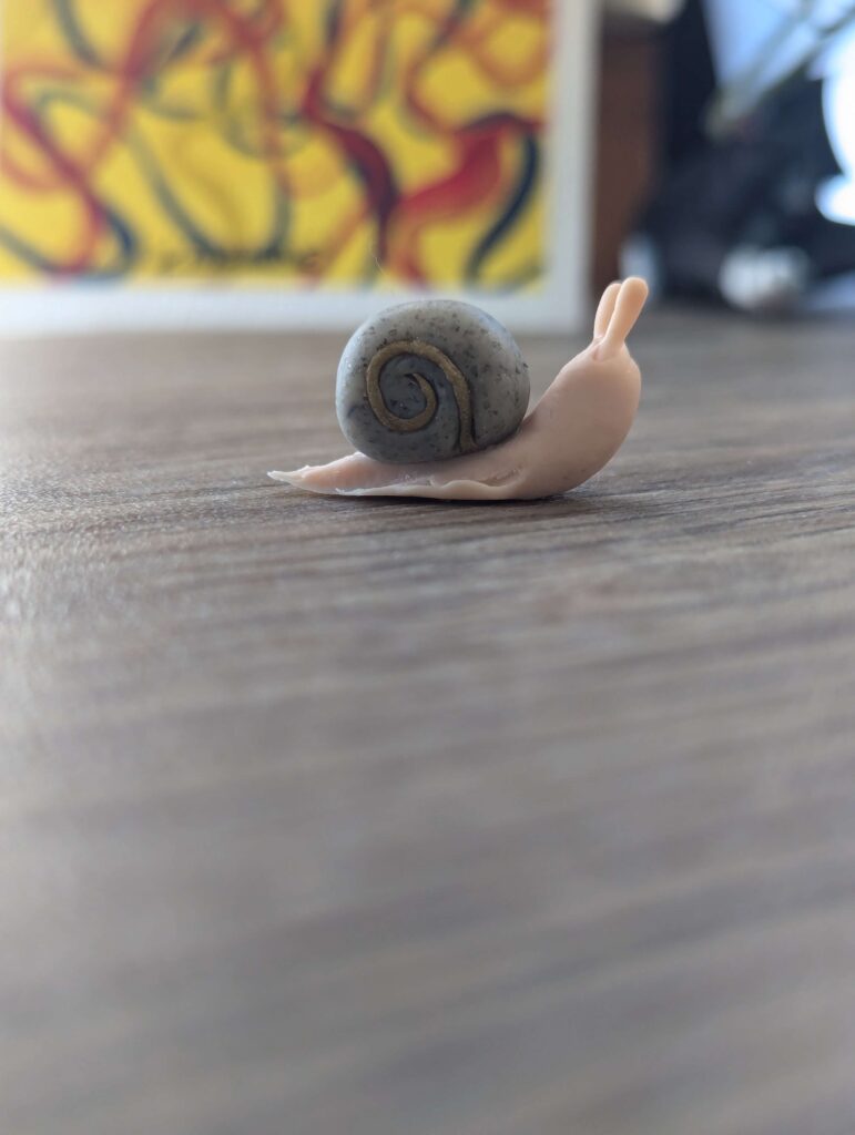 A tiny snail made from polymer clay. The shell is made of a gray granite color and the foot of the snail is a beige clay.