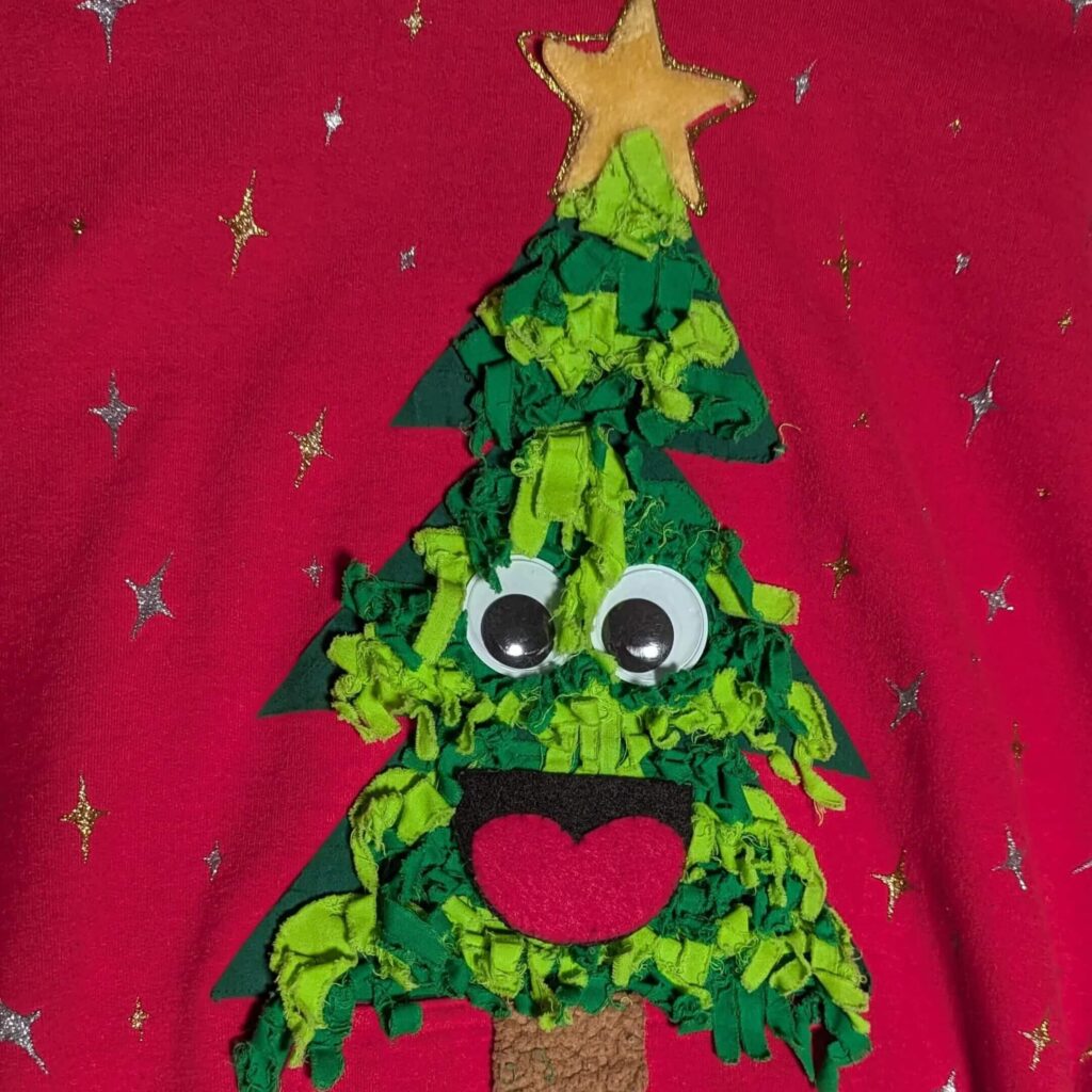 A red sweatshirt with a green christmas tree. The tree is made up of light and dark green fabric that has been cut into fringe and then sewn in a tree shape. There is a yellow fuzzy star at the top. The whole thing is surrounded by gold and silver puff paint stars.