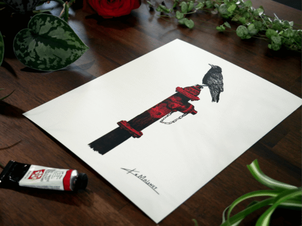 A close up angle shot of the crow on a hydrate art print. Which is an 8x10 art print of a crow sitting on a hydrate, the print is surrounded by Green plants as a flay lay frame and mixed into the plants are two red roses and a tube of red paint.