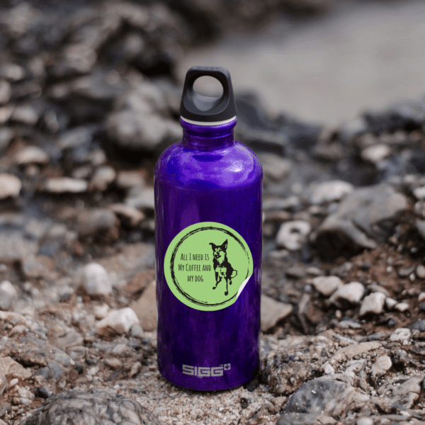 Purple water bottle with green circular sticker featuring 'All I Need Is My Coffee and My Dog' text and dog silhouette, photographed on rocky ground