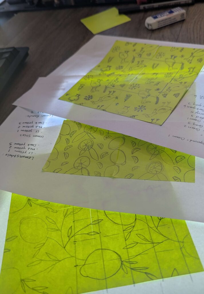 Hand-drawn surface pattern sketches on yellow post it notes, featuring repeating motifs of lemons, citrus slices, leaves, and birds, laid out on a desk with notes and tools nearby.