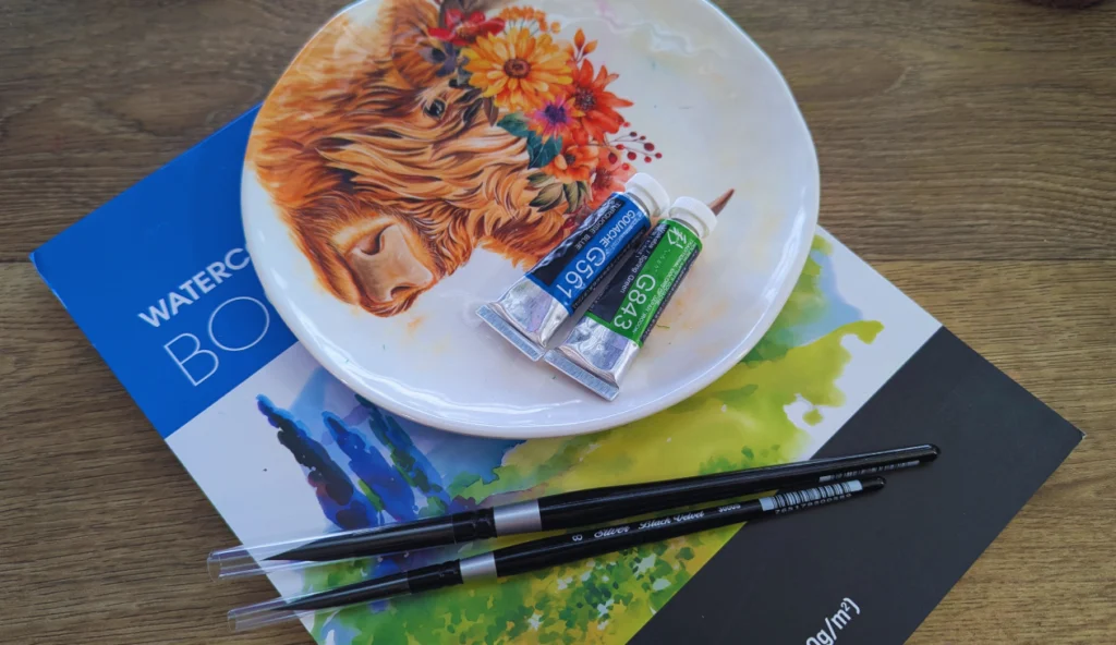 A worktable with art supplies stacked on it. There is a watercolor sketchbook on the bottom, with two black paint brushes laying on top. Also on the sketchbook is a plate with an image of a highland cow wearing a floral crown. On the plate are two tubes of gouache paint.